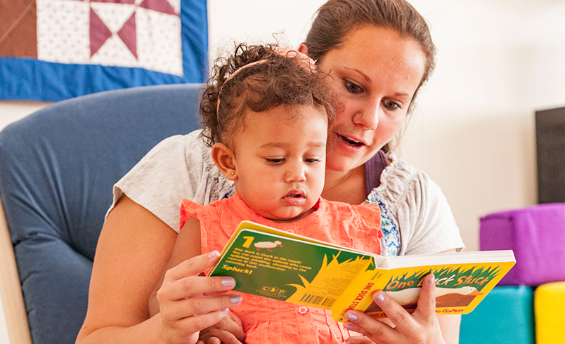 A nursery teacher is reading a picture book to a toddler girl