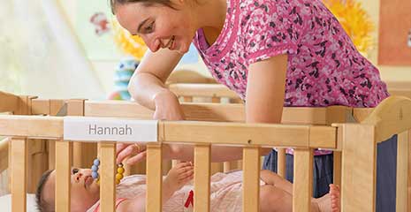 A nursery teacher plays with a baby in a cot.