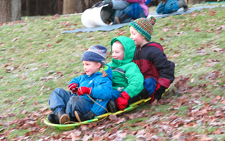 Three children bundled in winter gear sledding down a frosty hill covered in leaves.