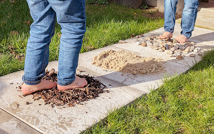 Two boys wearing jeans and walking barefoot along a sensory path layed with woodchips and pebbles.