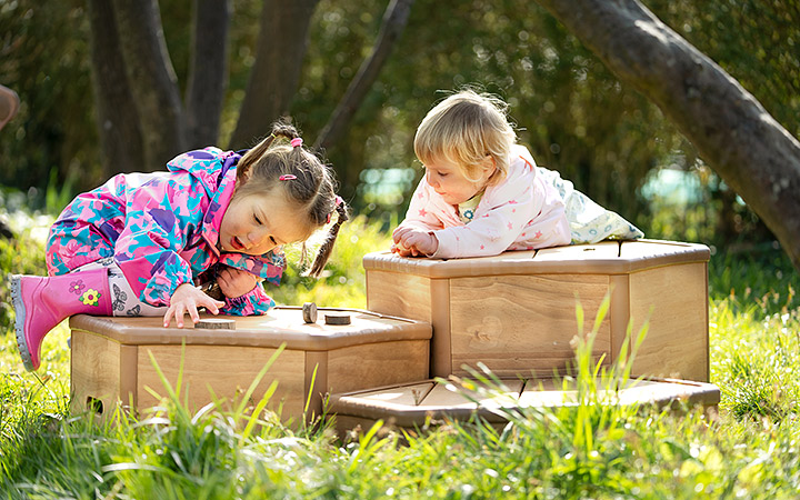 Two young children playing outdoors on wooden sensory boxes. One child, wearing a pink and blue raincoat, leans over to interact with the box, while the other, dressed in a white outfit, watches curiously. Lush greenery and trees surround them.