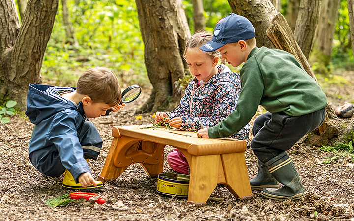 Three children exploring nature in a wooded area. One child examines items on a small wooden table using a magnifying glass, while the other two observe and participate in the discovery. They are dressed in warm, weather-appropriate clothing.