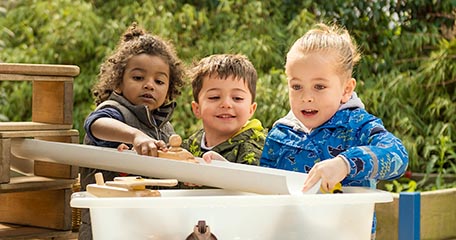 Three young children gathered around a water play station, experimenting with pouring and directing water. They appear engaged and excited, using ramps and tubs to explore water flow and movement.