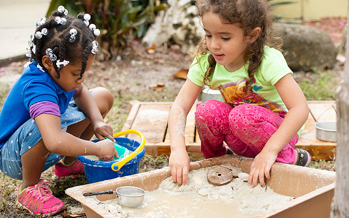 Two young girls engaged in sensory play with a mud and water mixture. One girl is using a blue bucket, while the other manipulates the mud with her hands. The outdoor environment is rich in natural play elements.