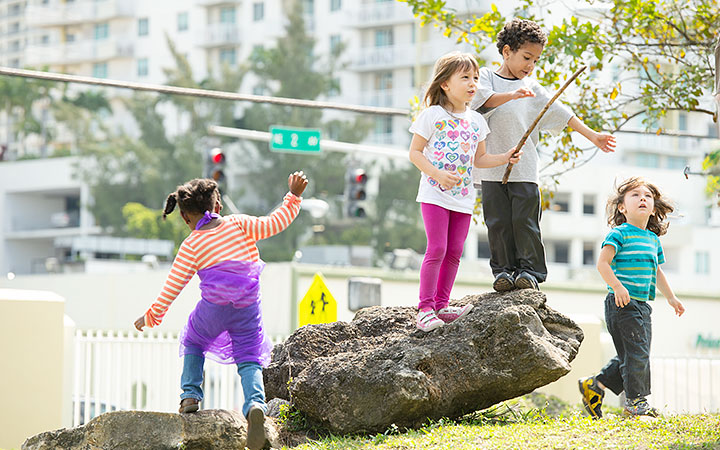 A group of four children playing on large rocks in an urban park setting. They are engaging in imaginative play, with one child holding a stick and others balancing on the rocks. The background features buildings, traffic lights, and trees.