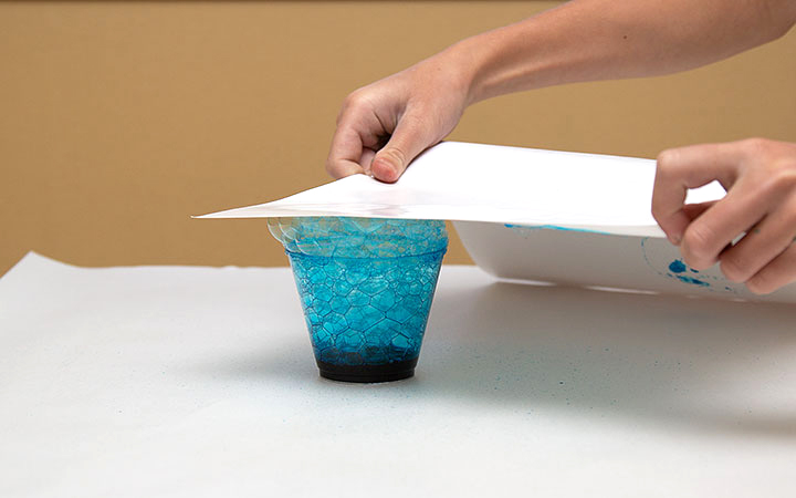 a woman’s hands are holding a sheet of paper onto a cup overflowing with blue bubbles