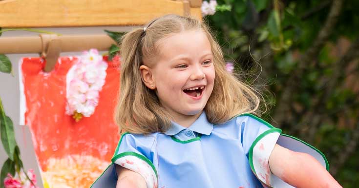 A girl laughing with her finished painting on an easel behind her.
