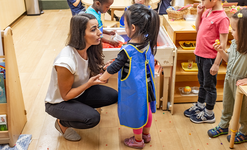 A nursery teacher is speaking to and gently holding the hands of a girl who is wearing a blue craft apron.