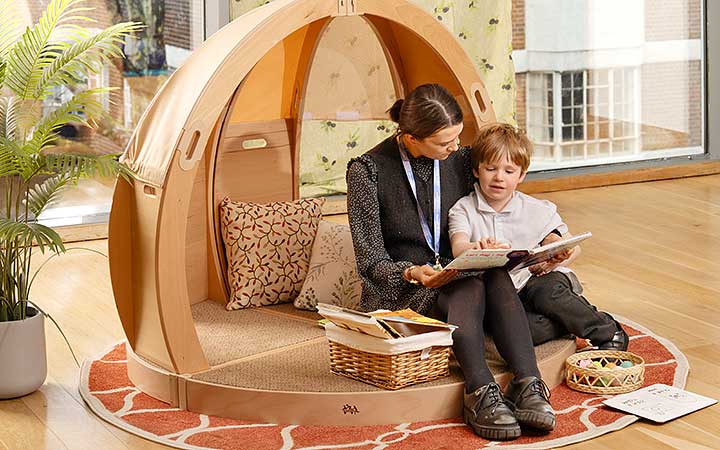 A primary aged boy with his teacher co-regulating in a calm corner made of solid wood and natural toned fabric.