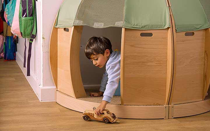 A toddler aged boy playing with a wooden vehicle in a calm corner made of solid wood and natural coloured fabric.