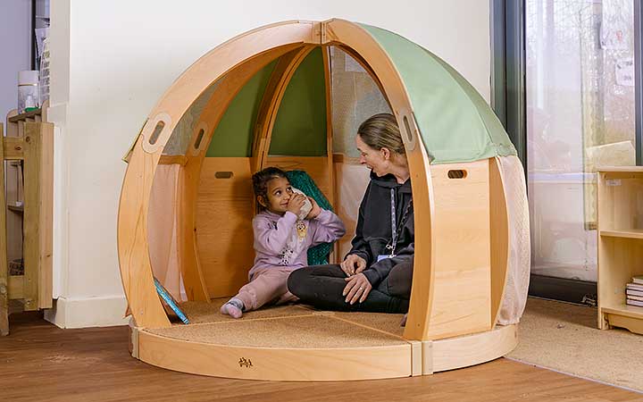 A nursery aged girl and her key person exploring a sea shell together in a solid wood den or regulation space.