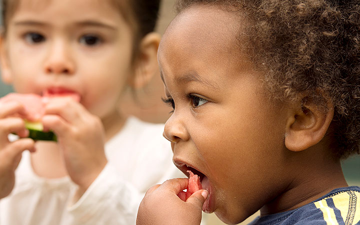 A close-up of two young children eating watermelon outdoors. The boy with curly hair is in focus as he takes a bite, while the girl in a white shirt is slightly blurred in the background, also enjoying her slice.