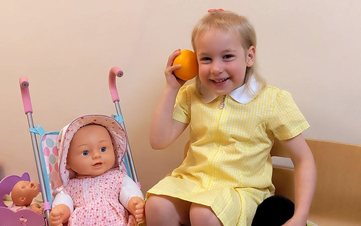 A young girl in a yellow dress sits indoors, playfully holding an orange to her ear as if listening to it. A baby doll in a stroller is beside her.