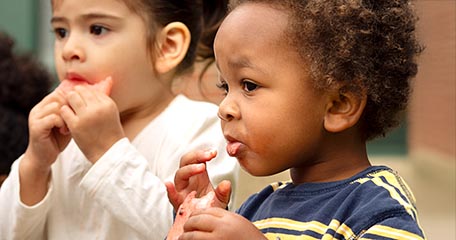 A zoomed-in shot of two toddlers enjoying watermelon outdoors. A boy with curly hair is in focus, eating a slice, while a girl in a white shirt is slightly blurred in the background, also taking a bite.