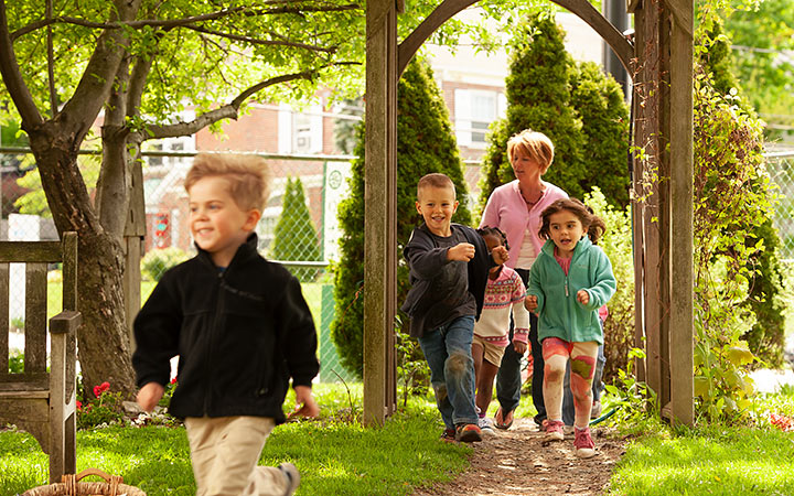Several yound children are running through a gate and along a path, followed by a nursery teacher