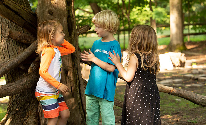 Three young children are standing by a tree and talking with each other