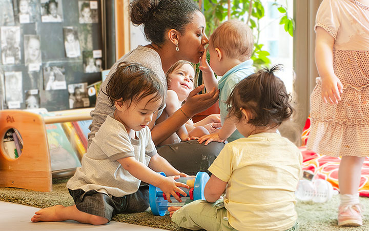 A group of four babies seated on the floor and interacting with eachother and their teacher
