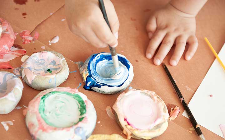 A child's hands holding a paintbrush and dipping it into a paint pot
