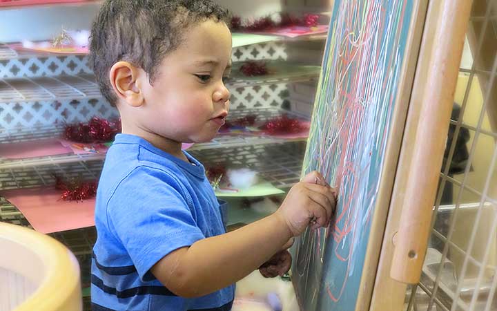 A small boy scribbling with chalk at a chalkboard mounted on a child-sized easel.