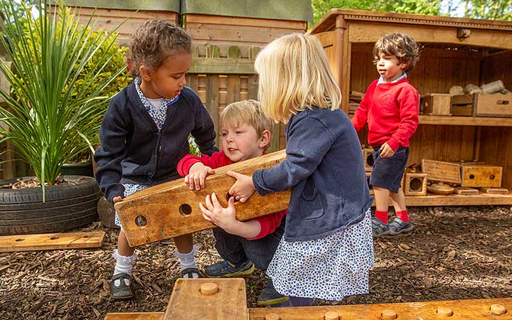 Three children helping eachother lift outdoor blocks in a school outdoors area