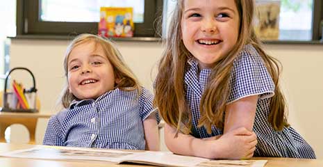 Two primary school girls leaning over a desk and laughing together