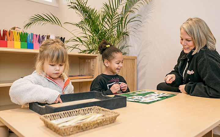 A teacher sits with two young children at a wooden table in a classroom. One child is engaged in a matching activity, while the other focuses on a black tray. A colorful set of pencils is visible on a shelf in the background.