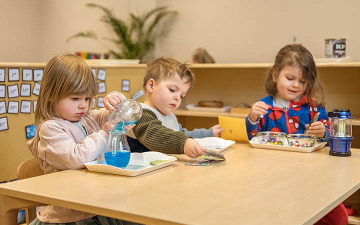 Three young children sit at a classroom table, participating in hands-on activities. One pours blue food coloured water from a beaker, another works with a small card, and the third child plays with colorful beads. The background features a learning environment with shelves and materials.