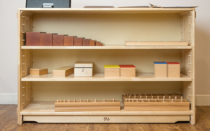 A wooden adjustable shelf stocked with various educational Montessori materials, including The Brown Stairs, Knobbed Cylinders, and the Binomial and Trinomial cube boxes.