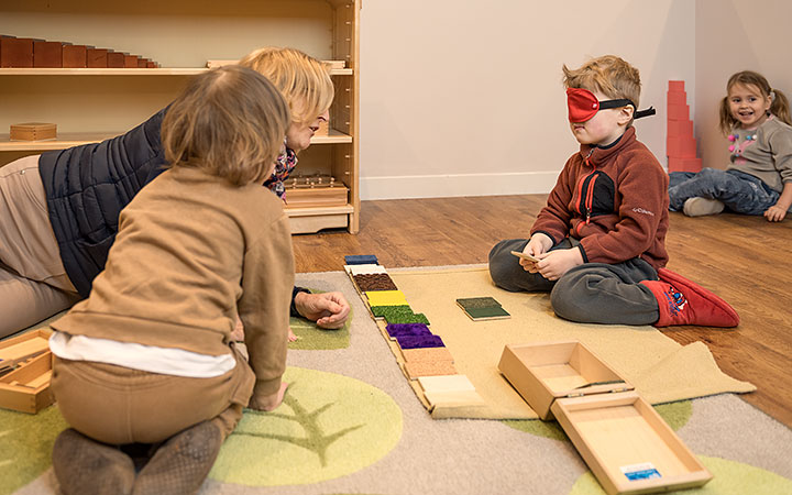 A young child wearing a blindfold sits on a classroom floor, engaged in a sensory matching activity with textured fabric swatches. Two other children and a teacher observe and participate in the activity.