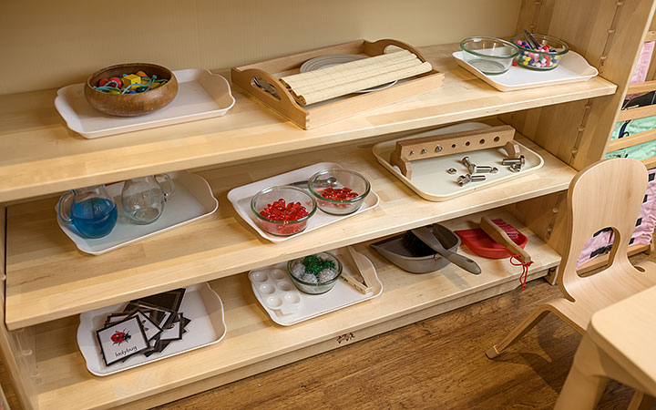 A Montessori classroom shelf displaying various practical life materials, including pouring sets, sorting trays, and fine motor skill activities, neatly arranged on white trays