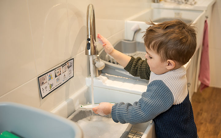 A young child wearing a sweater stands at a sink, washing dishes with soap and water. A visual instruction guide is posted on the wall next to the faucet.