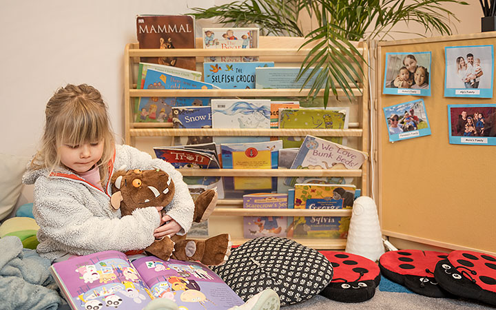A young girl sits in a cozy reading nook, holding a plush toy and flipping through a picture book. A wooden bookshelf filled with children's books is behind her, along with decorative cushions and family photos on the wall.