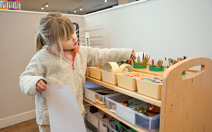 A young girl in a fuzzy jacket reaches for art supplies from a wooden organizer. She holds a piece of paper in one hand while selecting markers and pencils from neatly arranged bins.