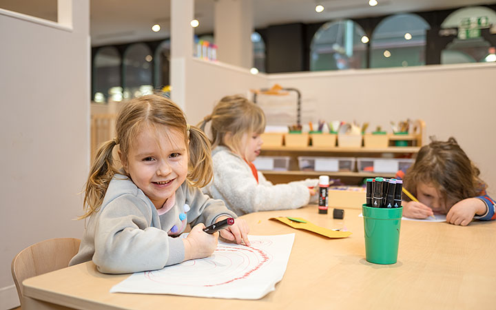 Three young children sit at a classroom table, drawing and coloring with markers. A girl in the foreground smiles while drawing, while the others focus on their artwork. The classroom environment is filled with creative supplies and learning materials.