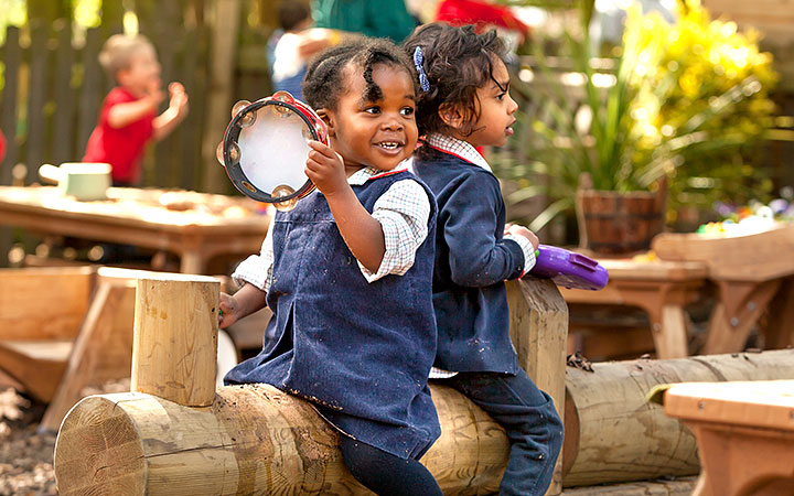 Two young children playing outdoors in a nursery setting. They sit on a wooden log, smiling and holding musical instruments.