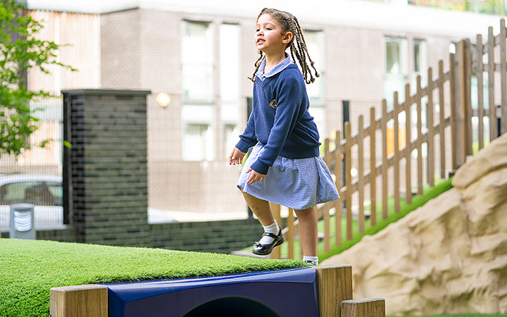 A reception-aged girl in school uniform climbing a grassy play structure in an outdoor learning environment. Behind her is a wooden fence and a brick building.