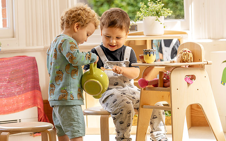 Two toddlers engaging in imaginative play indoors. One holds a cup while the other pours pretend tea out of a green pottery teapot. The classroom is bright and sunlit.