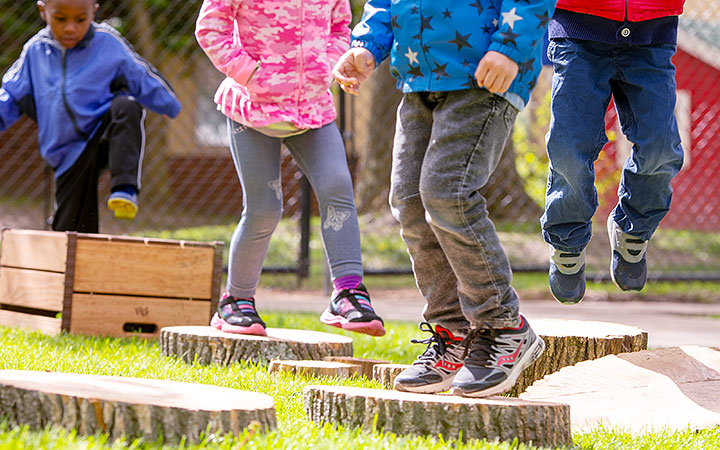 Children jumping between wooden stepping logs in an outdoor setting.