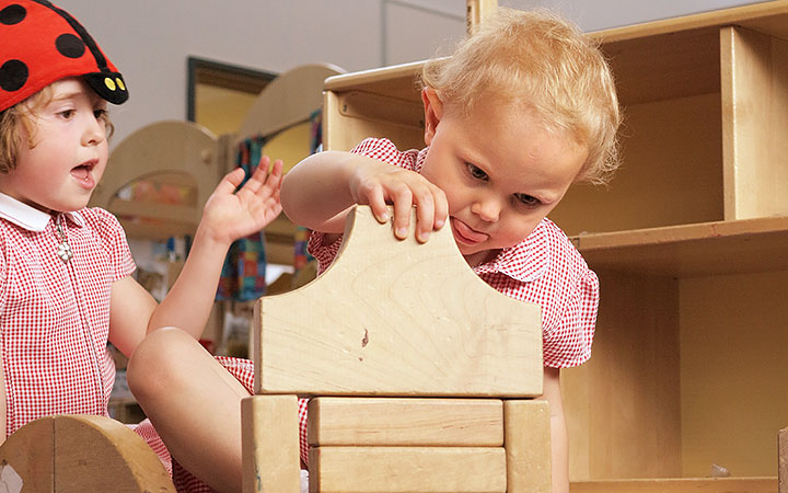 Two young reception-aged girls engaged in building a structure using wooden blocks. One child is carefully placing a block while the other watches with excitement.