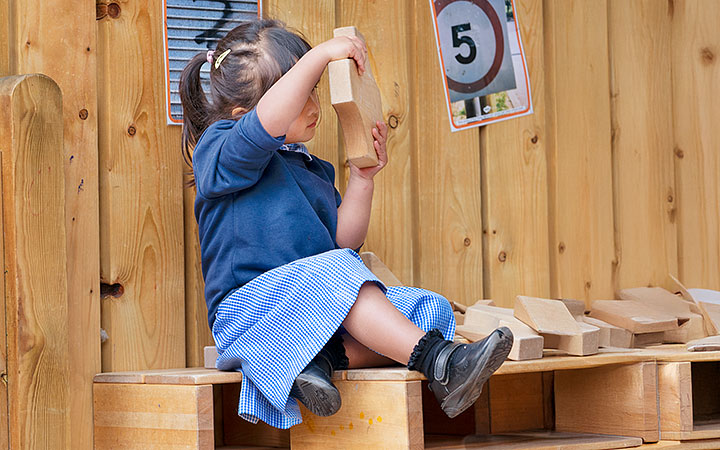 A young girl with dark hair tied in a ponytail, wearing a blue school cardigan and a blue checkered skirt, sitting on wooden blocks outdoors. She holds a wooden block up to her face, examining it. A wooden fence with a sign displaying the number '5' is visible in the background.