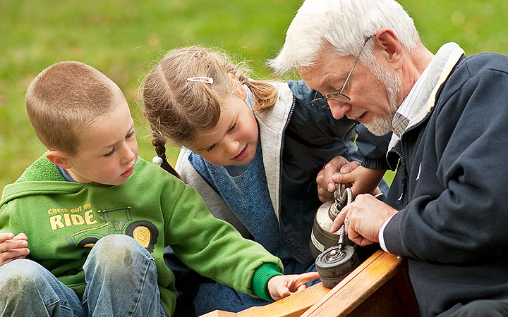 An elderly man with white hair and glasses shows two young children how to oil the wheels on their wooden toy truck. The children watch attentively as they learn hands-on skills.
