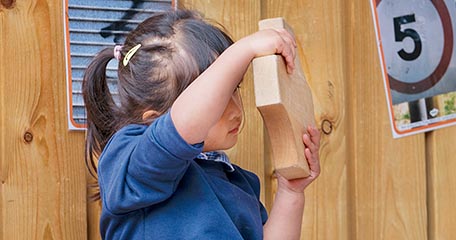 A close-up of a young girl in a blue school uniform closely examining a wooden block, holding it up to her face. A wooden fence with a number '5' sign is visible in the background.