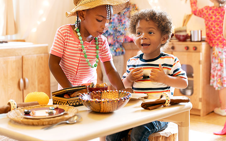 Two young children engage in imaginative play at a wooden table, set with woven baskets and natural materials. One child wears a straw hat and necklace while the other, in a striped shirt, smiles brightly, expressing excitement.
