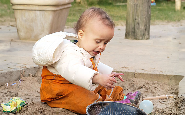 A baby in rust coloured waterproofs sitting in the sand pit and reaching for a toy