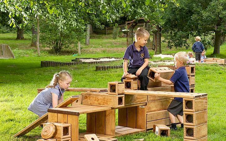 Children building with large wooden Outlast blocks on grass in an outdoor play area.