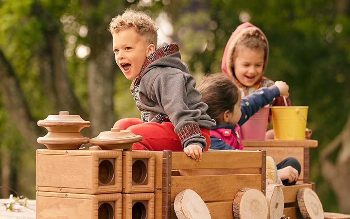 Three children playing outdoors and pretending to drive a wooden play vehicle built with Outlast blocks.