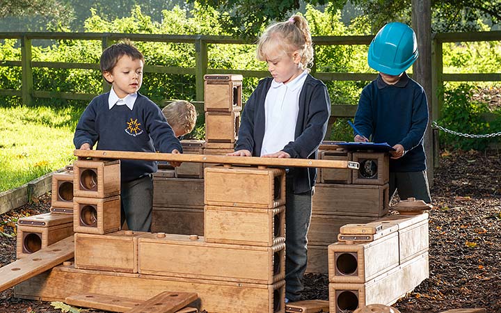 Children working together outdoors to build with wooden Outlast blocks. One child pretends to be a site manager and has a clipboard and safety helmet.