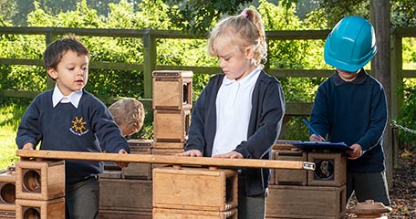 Children collaborating on a wooden block structure outdoors with one child taking notes.