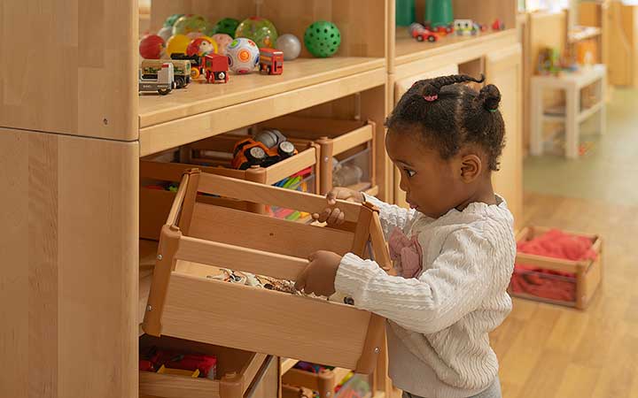 A girl putting a small wooden crate of toys back on a shelf.