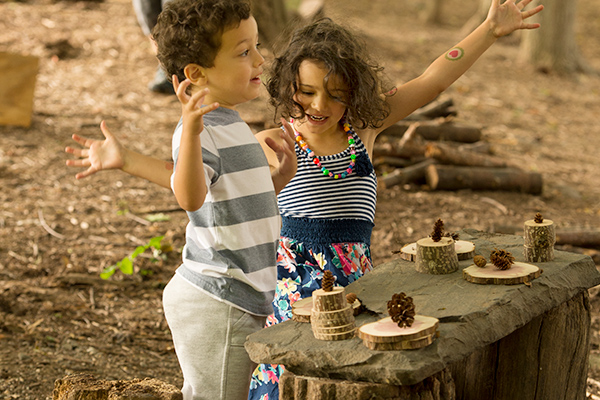 two children playing at a wooden bench with nature loose parts in the woods
